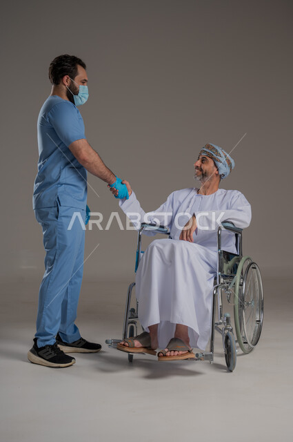  مفهوم العناية ورعاية المرضى والمعاقين، بورتريه لممرض يرتدي اليونيفورم والكمامة يقوم بالاعتناء برجل عربي خليجي عماني من ذوي الاحتياجات الخاصة يرتدي الدشداشة والعمامة يجلس على كرسي متحرك، ايماءات السرور والسعادة، خلفية رمادية