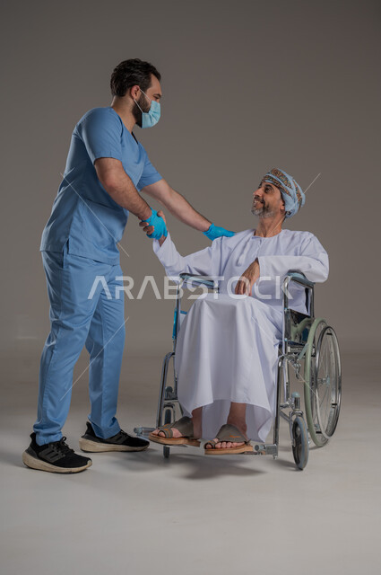  مفهوم العناية ورعاية المرضى والمعاقين، بورتريه لممرض يرتدي اليونيفورم والكمامة يقوم بالاعتناء برجل عربي خليجي عماني من ذوي الاحتياجات الخاصة يرتدي الدشداشة والعمامة يجلس على كرسي متحرك، ايماءات السرور والسعادة، خلفية رمادية