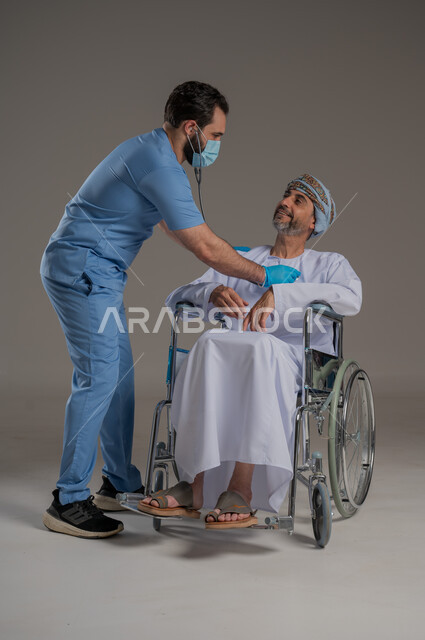  مفهوم العناية ورعاية المرضى والمعاقين، بورتريه لممرض يرتدي اليونيفورم والكمامة يقوم بالاعتناء برجل عربي خليجي عماني من ذوي الاحتياجات الخاصة يرتدي الدشداشة والعمامة يجلس على كرسي متحرك، ايماءات السرور والسعادة، خلفية رمادية