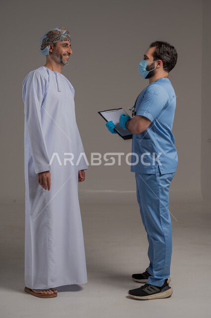 القيام بالرعاية الصحية , بورتريه مقرب لممرض يرتدي اليونيفورم يضع سماعة الأذن يقوم بالتأكد من سلامة رجل عربي خليجي عماني يرتدي الثوب التقليدي الدشداشة والعمامة وينظر للممرض بايماءات شكر وامتنان، مهن ووظائف طبية , خلفية رمادية 