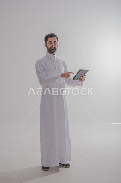 استخدام الأجهزة الحديثة والمتطورة، بورتريه لرجل عربي خليجي سعودي يرتدي الثوب التقليدي يحمل بيده جهاز التابليت، الدردشة مع الأهل والأصدقاء, تطبيقات وبرامج الأجهزة اللوحية، صورة الجسم بالطول الكامل, خلفية بيضاء 