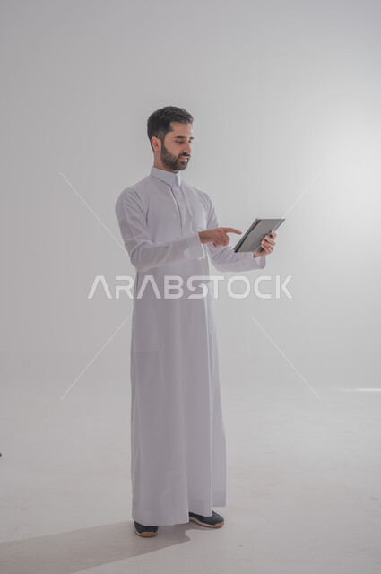 استخدام الأجهزة الحديثة والمتطورة، بورتريه لرجل عربي خليجي سعودي يرتدي الثوب التقليدي يحمل بيده جهاز التابليت، الدردشة مع الأهل والأصدقاء, تطبيقات وبرامج الأجهزة اللوحية، صورة الجسم بالطول الكامل, خلفية بيضاء 