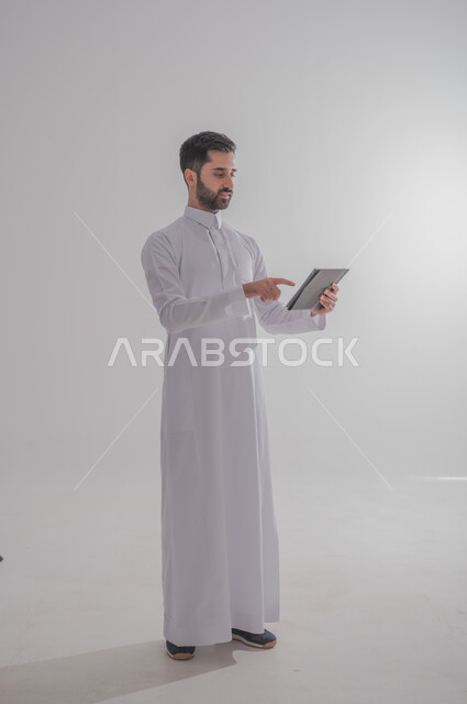 استخدام الأجهزة الحديثة والمتطورة، بورتريه لرجل عربي خليجي سعودي يرتدي الثوب التقليدي يحمل بيده جهاز التابليت، الدردشة مع الأهل والأصدقاء, تطبيقات وبرامج الأجهزة اللوحية، صورة الجسم بالطول الكامل, خلفية بيضاء 