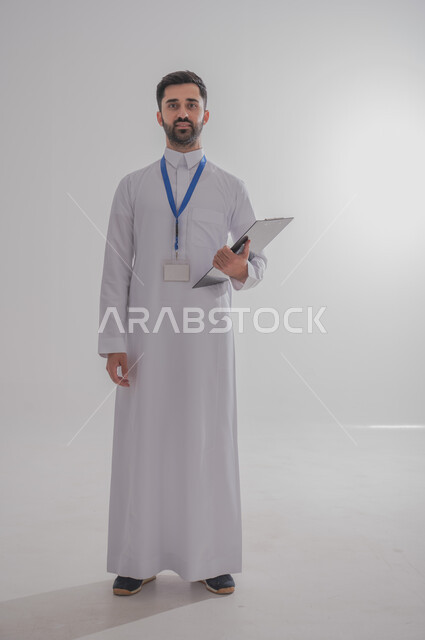 النظر إلى الكاميرا بايماءات الثقة بالنفس، مهن ووظائف مكتبية في المملكة العربية السعودية، بورتريه مقرب لرجل أعمال عربي سعودي خليجي مبتسم يرتدي الثوب التقليدي وبطاقة هوية تعريفية خاصة بالعمل يمسك بيده سنادة ورق سوداء, صورة الجسم بالطول الكامل، خلفية بيضاء