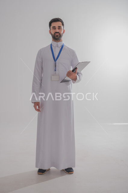 النظر إلى الكاميرا بايماءات الثقة بالنفس، مهن ووظائف مكتبية في المملكة العربية السعودية، بورتريه مقرب لرجل أعمال عربي سعودي خليجي مبتسم يرتدي الثوب التقليدي وبطاقة هوية تعريفية خاصة بالعمل يمسك بيده سنادة ورق سوداء, صورة الجسم بالطول الكامل، خلفية بيضاء
