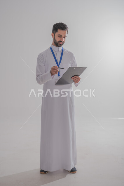 تدوين المعلومات و الملاحظات المهمة، مهن ووظائف مكتبية في المملكة العربية السعودية، بورتريه مقرب لرجل أعمال عربي سعودي خليجي يرتدي الثوب التقليدي وبطاقة هوية تعريفية خاصة بالعمل يمسك بيده سنادة ورق سوداء, صورة الجسم بالطول الكامل، خلفية بيضاء