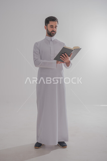 انشطة وفعاليات ترفيهية طلابي، مفهوم التمعن والتركيز، قضاء الوقت بالقراءة والتعلم والثقافة، بورتريه لرجل عربي سعودي خليجي يرتدي الثوب التقليدي يحمل بيديه كتاب، الجسم بالطول الكامل، خلفية بيضاء