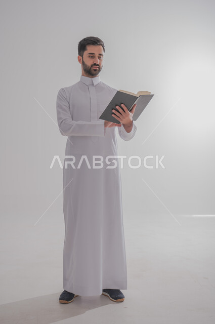انشطة وفعاليات ترفيهية طلابي، مفهوم التمعن والتركيز، قضاء الوقت بالقراءة والتعلم والثقافة، بورتريه لرجل عربي سعودي خليجي يرتدي الثوب التقليدي يحمل بيديه كتاب، الجسم بالطول الكامل، خلفية بيضاء