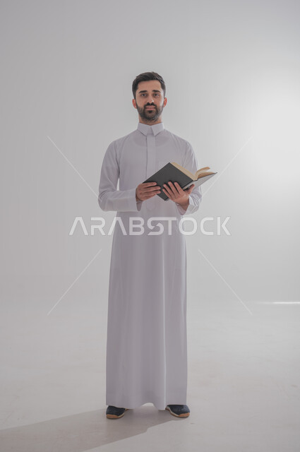 النظر إلى الكاميرا بايماءات الفرح والسرور، انشطة وفعاليات ترفيهية طلابي، مفهوم التمعن والتركيز، قضاء الوقت بالقراءة والتعلم والثقافة، بورتريه لرجل عربي سعودي خليجي يرتدي الثوب التقليدي يحمل بيديه كتاب، الجسم بالطول الكامل، خلفية بيضاء