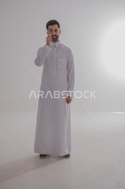 الفحص والاستكشاف والرؤية عن قرب، مفهوم توضيح الصورة، ايماءات تدل على السعادة والسرور، بورتريه لرجل عربي خليجي سعودي يرتدي الثوب التقليدي وينظر من خلال العدسة المكبرة، التدقيق والتركيز باستخدام الأدوات البصرية، الجسم بالطول الكامل، خلفية رمادية