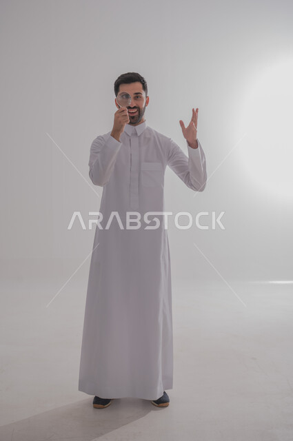 ايماءات الفرح والسرور، الفحص والرؤية عن قرب، مفهوم توضيح الصورة، ايماءات تدل على السعادة والسرور، بورتريه لرجل خليجي سعودي يرتدي الثوب التقليدي وينظر من خلال العدسة المكبرة بحماس، التدقيق والتركيز باستخدام الأدوات البصرية، الجسم بالطول الكامل، خلفية رمادية