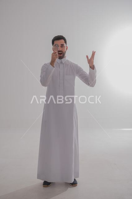 ايماءات الفرح والسرور، الفحص والرؤية عن قرب، مفهوم توضيح الصورة، ايماءات تدل على السعادة والسرور، بورتريه لرجل خليجي سعودي يرتدي الثوب التقليدي وينظر من خلال العدسة المكبرة بتفاجؤ وذهول، التدقيق والتركيز باستخدام الأدوات البصرية، الجسم بالطول الكامل، خلفية رمادية