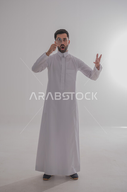 ايماءات الفرح والسرور، الفحص والرؤية عن قرب، مفهوم توضيح الصورة، ايماءات تدل على السعادة والسرور، بورتريه لرجل خليجي سعودي يرتدي الثوب التقليدي وينظر من خلال العدسة المكبرة بتفاجؤ وذهول، التدقيق والتركيز باستخدام الأدوات البصرية، الجسم بالطول الكامل، خلفية رمادية