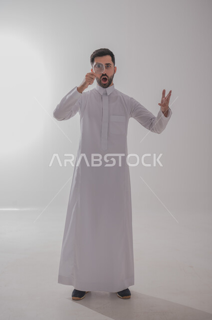 ايماءات الفرح والسرور، الفحص والرؤية عن قرب، مفهوم توضيح الصورة، ايماءات تدل على السعادة والسرور، بورتريه لرجل خليجي سعودي يرتدي الثوب التقليدي وينظر من خلال العدسة المكبرة بتفاجؤ وذهول، التدقيق والتركيز باستخدام الأدوات البصرية، الجسم بالطول الكامل، خلفية رمادية