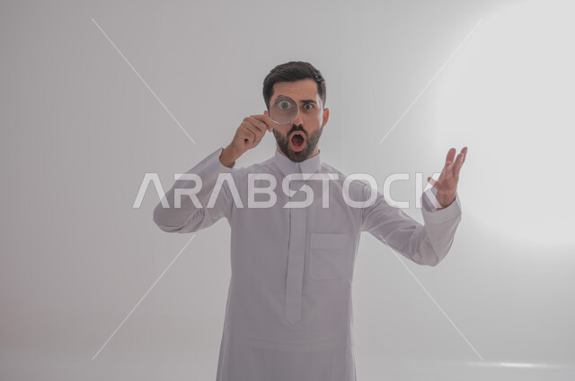 مفهوم توضيح الصورة، ايماءات الفرح والسرور، الفحص والرؤية عن قرب، ايماءات تدل على السعادة والسرور، بورتريه مقرب لرجل خليجي سعودي يرتدي الثوب التقليدي وينظر من خلال العدسة المكبرة بتفاجؤ وذهول، التدقيق والتركيز باستخدام الأدوات البصرية، خلفية رمادية