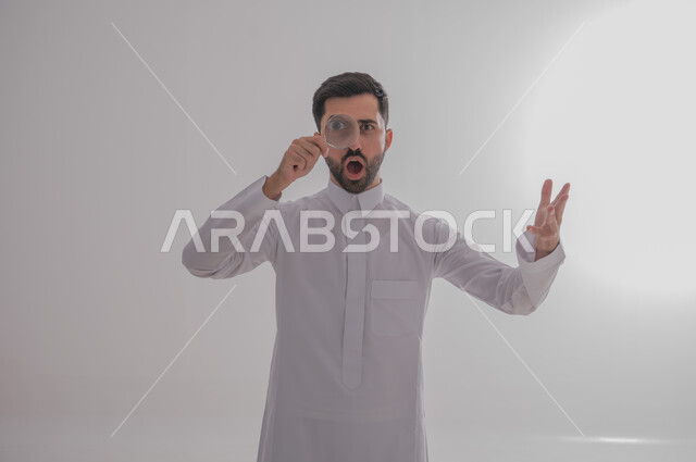مفهوم توضيح الصورة، ايماءات الفرح والسرور، الفحص والرؤية عن قرب، ايماءات تدل على السعادة والسرور، بورتريه مقرب لرجل خليجي سعودي يرتدي الثوب التقليدي وينظر من خلال العدسة المكبرة بتفاجؤ وذهول، التدقيق والتركيز باستخدام الأدوات البصرية، خلفية رمادية