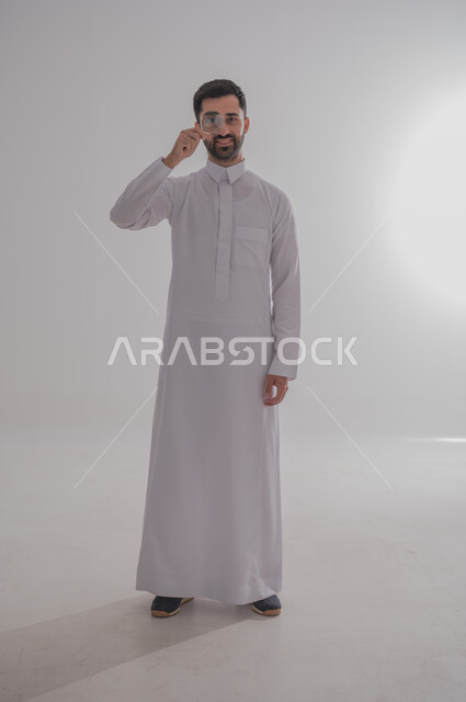 الفحص والاستكشاف والرؤية عن قرب، مفهوم توضيح الصورة، ايماءات تدل على السعادة والسرور، بورتريه لرجل عربي خليجي سعودي يرتدي الثوب التقليدي وينظر من خلال العدسة المكبرة، التدقيق والتركيز باستخدام الأدوات البصرية، الجسم بالطول الكامل، خلفية رمادية