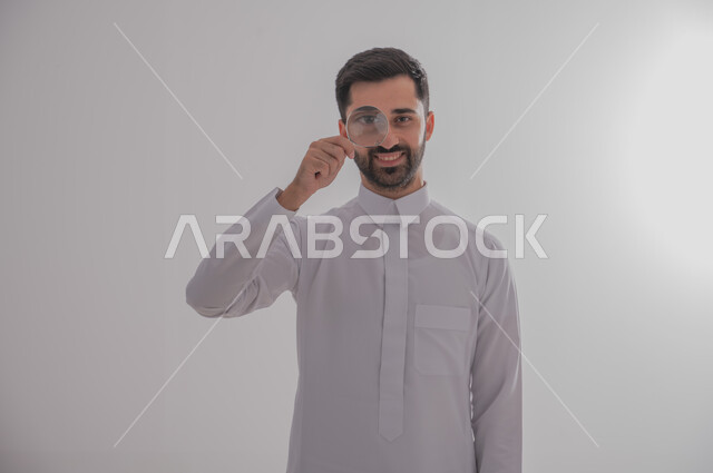التدقيق والتركيز باستخدام الأدوات البصرية، الفحص والاستكشاف والرؤية عن قرب، مفهوم توضيح الصورة، ايماءات تدل على السعادة والسرور، بورتريه مقرب لرجل عربي خليجي سعودي يرتدي الثوب التقليدي وينظر من خلال العدسة المكبرة، خلفية رمادية
