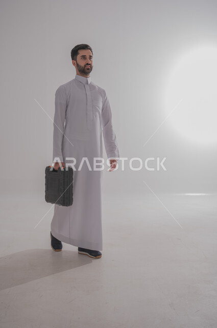 الذهاب للعمل بنشاط وحيوية، ورش عمل وتصليح في المملكة العربية السعودية، تصليح و صيانة السيارات، بورتريه لرجل عربي خليجي سعودي يرتدي الثوب التقليدي يحمل حقيبة معدات مغلقة، صورة الجسم بالطول الكامل، خلفية بيضاء