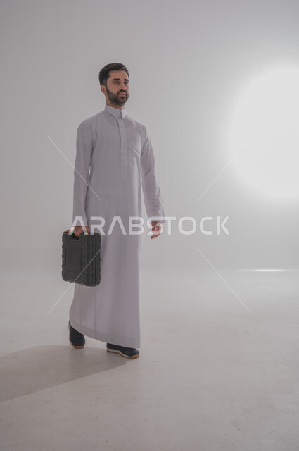 الذهاب للعمل بنشاط وحيوية، ورش عمل وتصليح في المملكة العربية السعودية، تصليح و صيانة السيارات، بورتريه لرجل عربي خليجي سعودي يرتدي الثوب التقليدي يحمل حقيبة معدات مغلقة، صورة الجسم بالطول الكامل، خلفية بيضاء