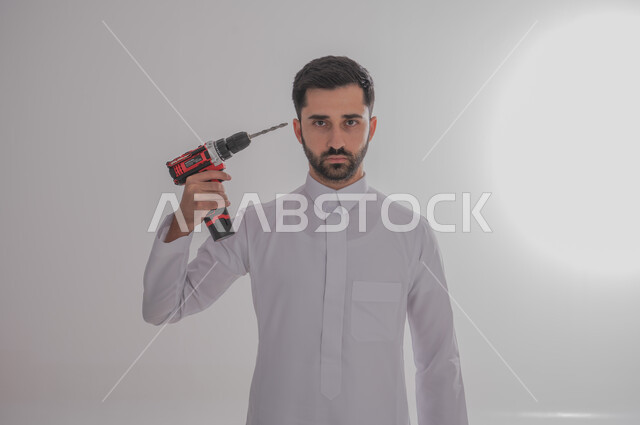 استخدام أدوات ومعدات الصيانة لاجراء التصليحات، المهارة و اتقان العمل، بورتريه لرجل عربي سعودي خليجي يرتدي الثوب التقليدي ينظر إلى الكاميرا بإيماءات الغضب يحمل بيده فرد ثاقب يوجهه نحو رأسه، خلفية بيضاء