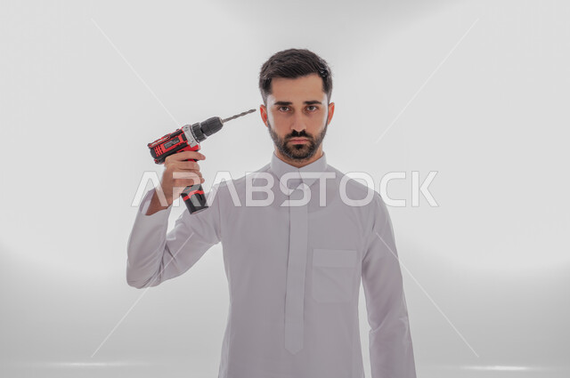 Using maintenance tools and equipment to make repairs, skill and mastery of work, portrait of a Saudi Arabian Gulf man wearing traditional thobe looking at the camera with angry gestures holding a piercing saw in his hand and pointing it towards his head, white background
