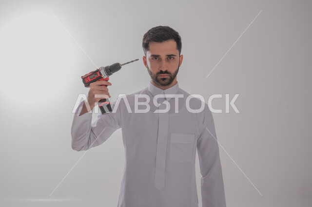 استخدام أدوات ومعدات الصيانة لاجراء التصليحات، المهارة و اتقان العمل، بورتريه لرجل عربي سعودي خليجي يرتدي الثوب التقليدي ينظر إلى الكاميرا بإيماءات الغضب يحمل بيده فرد ثاقب يوجهه نحو رأسه، خلفية بيضاء