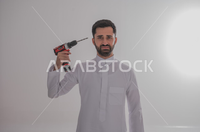 Using maintenance tools and equipment to make repairs, skill and mastery of work, portrait of a Saudi Arabian Gulf man wearing traditional thobe looking at the camera with crying gestures holding a piercing saw in his hand and pointing it towards his head, white background