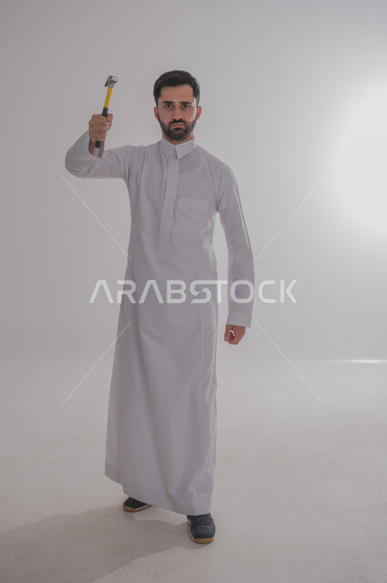 Skill and mastery of work, using maintenance tools and equipment to make repairs, portrait of a Saudi Arabian Gulf man wearing traditional thobe looking at the camera with angry gestures holding a hammer in his hand, full body, white background