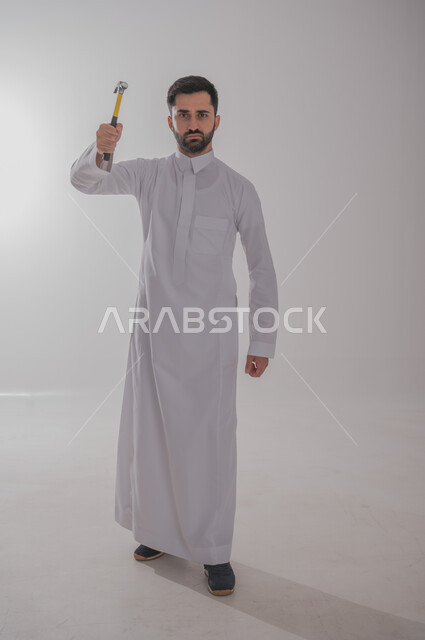 Skill and mastery of work, using maintenance tools and equipment to make repairs, portrait of a Saudi Arabian Gulf man wearing traditional thobe looking at the camera with angry gestures holding a hammer in his hand, full body, white background