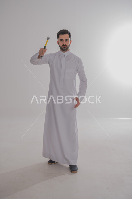 المهارة و اتقان العمل، استخدام أدوات ومعدات الصيانة لاجراء التصليحات، بورتريه لرجل عربي سعودي خليجي يرتدي الثوب التقليدي ينظر إلى الكاميرا بإيماءات الغضب يحمل بيده مطرقة، الجسم بالطول الكامل، خلفية بيضاء