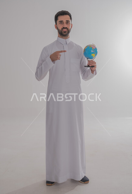 Pointing to a specific place on the map, searching and exploring around the world, portrait of a smiling Saudi Arabian Gulf man wearing traditional thobe holding a globe, concept of travel and tours, full body, white background