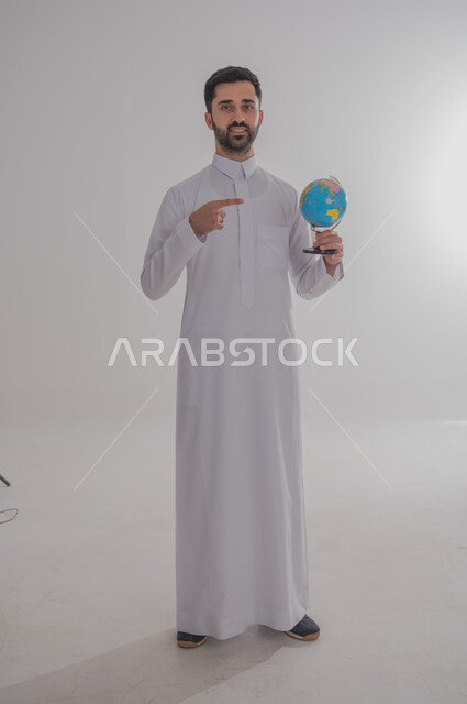 الاشارة إلى مكان معين على الخريطة, البحث والاستكشاف حول العالم, بورتريه لرجل عربي خليجي سعودي مبتسم يرتدي الثوب التقليدي يحمل بيده مجسم الكرة الارضية, مفهوم الرحلات والجولات السياحية, الجسم بالطول الكامل، خلفية بيضاء
