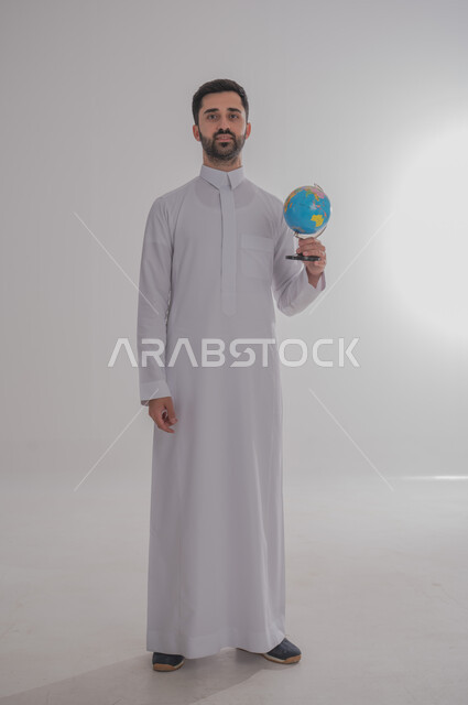 Locating Arab countries and states on the world map, research and exploration around the universe, portrait of a smiling Saudi Arabian Gulf man wearing traditional thobe holding a globe in his hand, concept of travel and tours, full body, white background