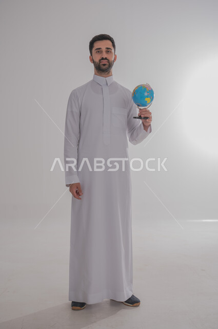تحديد موقع الدول والبلدان العربية على خريطة العالم, البحث والاستكشاف حول الكون, بورتريه لرجل عربي خليجي سعودي مبتسم يرتدي الثوب التقليدي يحمل بيده مجسم الكرة الارضية, مفهوم الرحلات والجولات السياحية, الجسم بالطول الكامل، خلفية بيضاء 