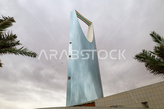 التصميم المتطور والحديث للأبراج وناطحات السحاب في السعودية، أماكن سياحية مشهورة, الفن الهندسي المعماري للمنشآت التجارية في مدينة الرياض, صورة مقربة من الأسفل لمبنى برج المملكة في وقت النهار, منظر السماء الممتلئة بالغيوم