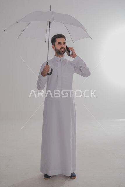 سهولة إجراء مكالمة هاتفية تحت المطر، التظلل من أشعة الشمس، الحماية من الأمطار في الشتاء، بورتريه لرجل عربي خليجي سعودي يرتدي الثوب التقليدي  يحمل بيده هاتف محمول يقوم بإجراء مكالمة هاتفية، صورة لكامل الجسم، خلفية بيضاء 
