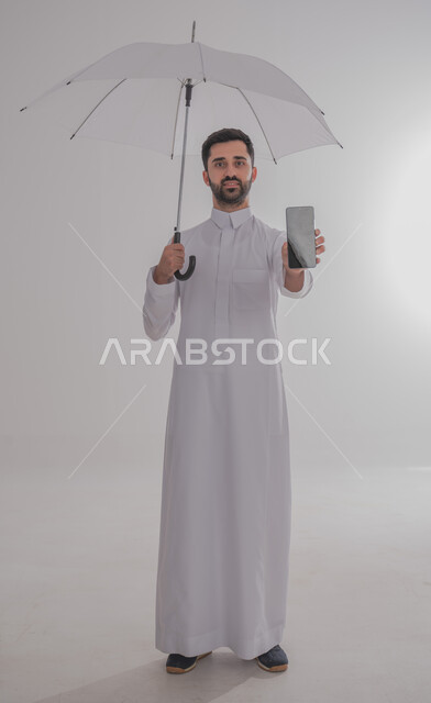 استخدام التقنيات الحديثة والأجهزة المتطورة، التظلل من أشعة الشمس، الحماية من الأمطار في الشتاء، بورتريه لرجل عربي خليجي سعودي يرتدي الثوب التقليدي  يحمل بيده هاتف محمول يقوم بعرض شاشة سوداء فارغة، صورة لكامل الجسم، خلفية بيضاء 