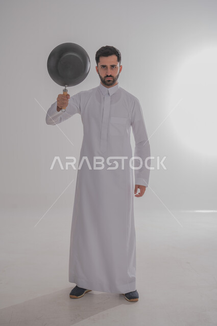 ايماءات وجه تدل على الغضب والتعصيب، عدم السيطرة على النفس عند الانزعاج، بورتريه لرجل عربي خليجي سعودي غاضب يرتدي الثوب التقليدي يحمل بيده مقلاة يرفعها للأعلى، الجسم بالطول الكامل، خلفية بيضاء 
