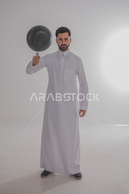 ايماءات وجه تدل على الغضب والتعصيب، عدم السيطرة على النفس عند الانزعاج، بورتريه لرجل عربي خليجي سعودي غاضب يرتدي الثوب التقليدي يحمل بيده مقلاة يرفعها للأعلى، الجسم بالطول الكامل، خلفية بيضاء 