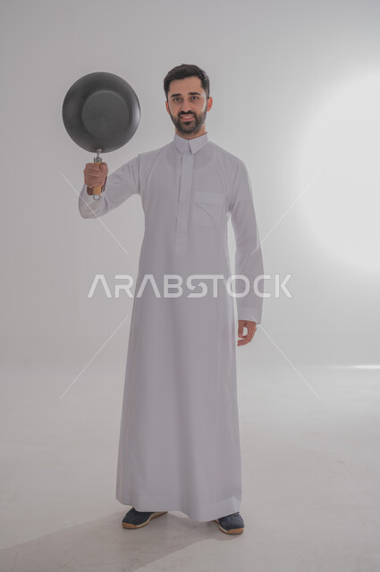 ايماءات وجه تدل على الغضب والتعصيب، عدم السيطرة على النفس عند الانزعاج، بورتريه لرجل عربي خليجي سعودي غاضب يرتدي الثوب التقليدي يحمل بيده مقلاة يرفعها للأعلى، الجسم بالطول الكامل، خلفية بيضاء 