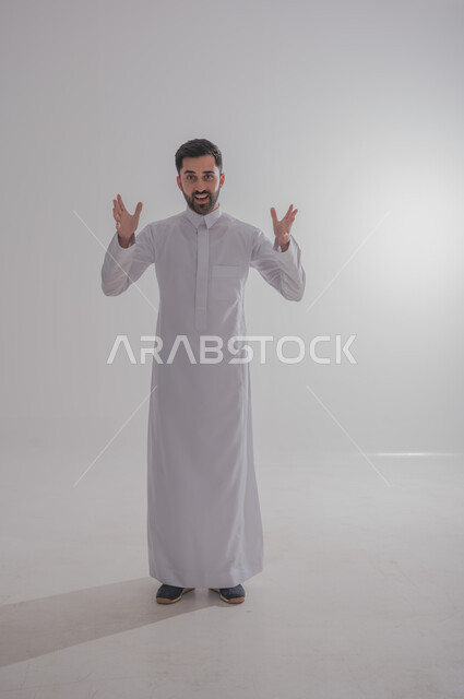 رفع اليدين في الهواء بتعابير الغضب، النظر للكاميرا بإيماءات الانزعاج وعدم الرضا، بورتريه لرجل عربي خليجي سعودي يرتدي الثوب التقليدي يرفع كفيه بتذمر وسخط، الجسم بالطول الكامل، خلفية بيضاء