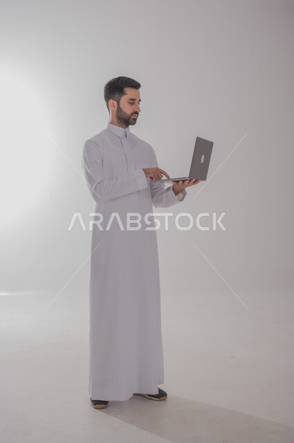 مفهوم العمل عن بعد، بورتريه لرجل عربي خليجي سعودي يحمل بيده جهاز الحاسوب المحمول، انجاز المهام المكتبية، تصفح الانترنت ووسائل التواصل الاجتماعي، احتراف وظائف الحاسوب وتكنولوجيا المعلومات، الجسم بالطول الكامل، خلفية بيضاء