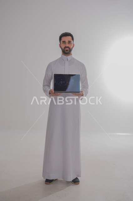 مفهوم العمل عن بعد، بورتريه لرجل عربي خليجي سعودي يحمل بيده جهاز الحاسوب المحمول، انجاز المهام المكتبية، تصفح الانترنت ووسائل التواصل الاجتماعي، احتراف وظائف الحاسوب وتكنولوجيا المعلومات، الجسم بالطول الكامل، خلفية بيضاء