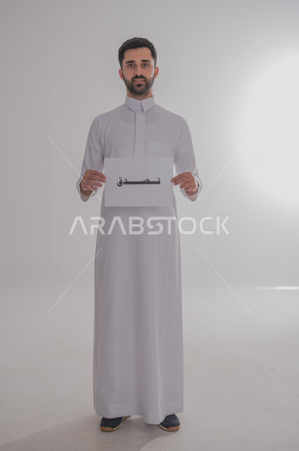 مفهوم طلب العون والمساعدة، لوحه بيضاء مربعه، استخدام لوحات توضيحية تعريفية، بورتريه لرجل عربي خليجي سعودي يرتدي الثوب التقليدي يحمل بيده موك اب مربع أبيض مكتوب عليه تصدق، ينظر إلى الكاميرا، صورة الجسم بالكامل، خلفية بيضاء  