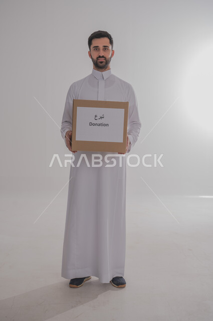 مفهوم طلب العون والمساعدة، لوحه بيضاء مربعه، استخدام لوحات توضيحية تعريفية، بورتريه لرجل عربي خليجي سعودي يرتدي الثوب التقليدي يحمل بيده صندوق مربع أبيض مكتوب عليه تبرع ينظر إلى الكاميرا، صورة الجسم بالكامل، خلفية بيضاء 