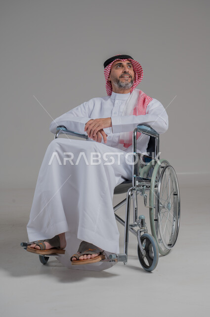 ضعف القدرة الحركية وصعوبة المشي، بورتريه لشاب عربي خليجي سعودي من ذوي الاحتياجات الخاصة يرتدي الثوب التقليدي والشماغ يجلس على كرسي متحرك على وجهه إيماءات البشاشة والسرور، الجسم بالطول الكامل، خلفية رمادية