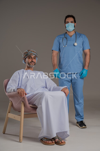 ايماءات السرور والسعادة، مفهوم العناية ورعاية المرضى والمسنين، بورتريه لممرض يرتدي اليونيفورم والكمامة يقوم بالاعتناء برجل عربي خليجي عماني مسن يرتدي الدشداشة والعمامة يجلس على كرسي ، خلفية رمادية