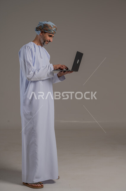 مفهوم تسهيل الاعمال والقدرة على إنجاز المهام عن بعد، استخدام التقنيات الحديثة والأجهزة المتطورة ، بورتريه لشاب عربي خليجي عماني يرتدي الدشداشة والعمامة يمسك بيده حاسوب محمول تظهر عليه علامات الفرح والسرور، الجسم بالطول الكامل، خلفية رمادية