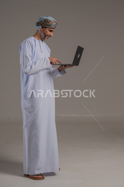 The concept of facilitating work and the ability to accomplish tasks remotely, using modern technologies and advanced devices, a portrait of a young Gulf Arab Omani man wearing a dishdasha and turban holding a laptop in his hand, showing signs of joy and happiness, full-length body, gray background